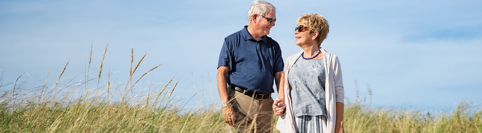 An older couple wearing sunglasses holds hands and walks through tall grass in a field on a clear day while looking at each other